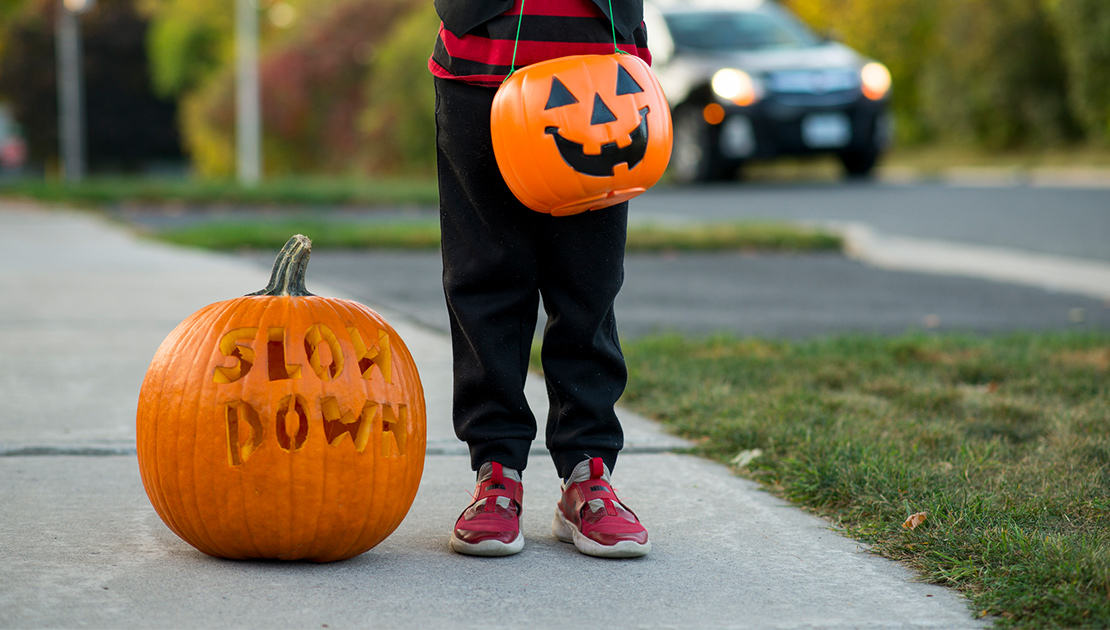 child holding jackolantern trick-or-treat basket with a pumpkin next to his feet. Pumpkin has words Slow Down carved into it